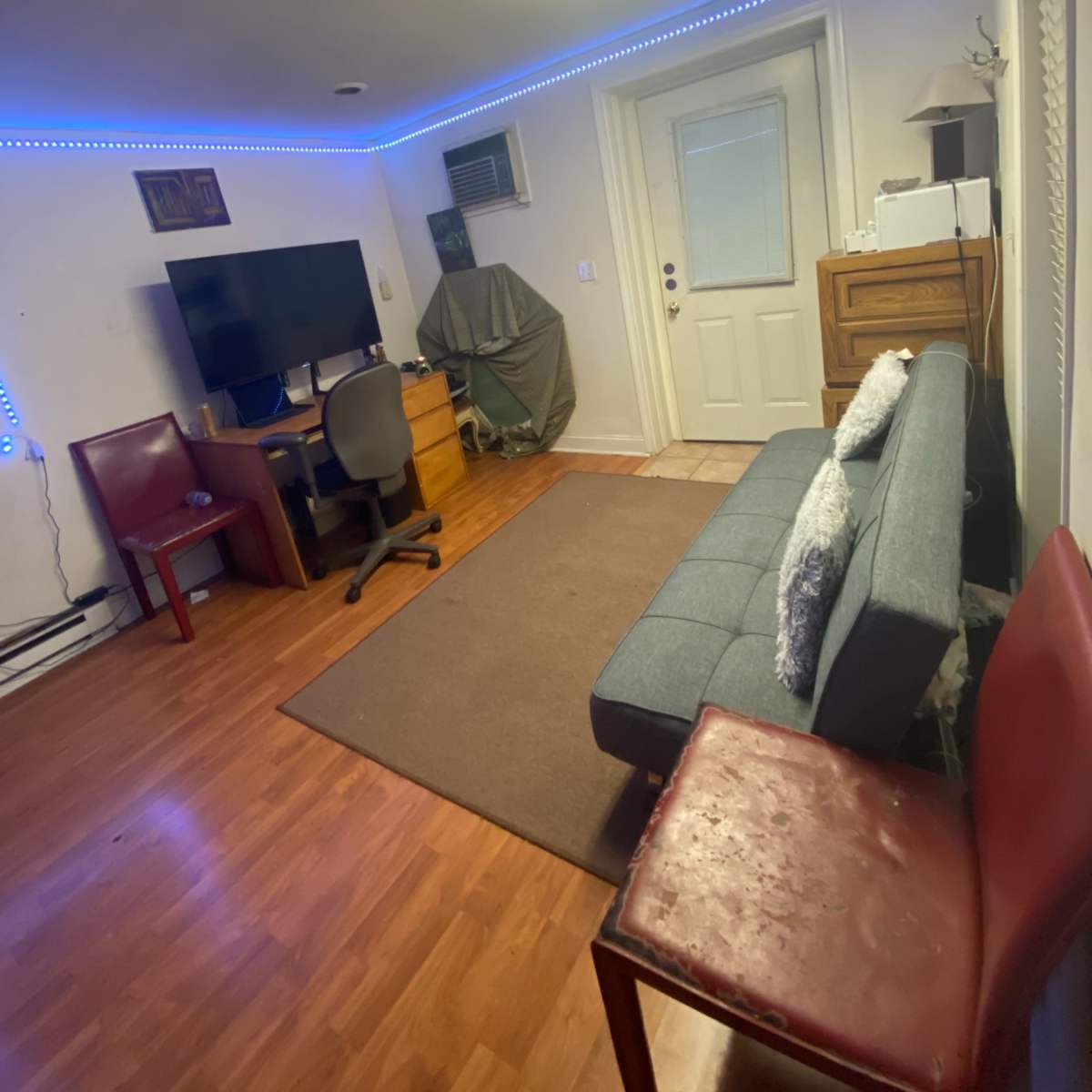 A small room with a gray sofa, a desk with a computer monitor, and two chairs, illuminated by blue LED light strips along the ceiling.