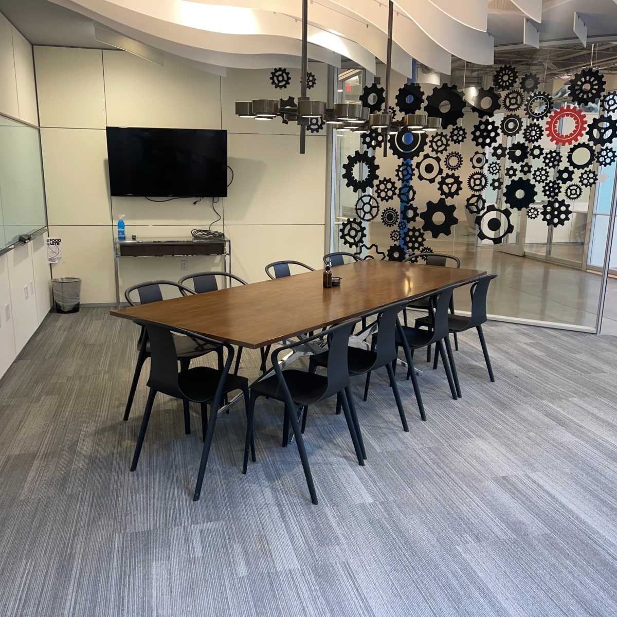 A meeting room features a long wooden table surrounded by black chairs, with a television on one wall and decorative gear shapes on another.
