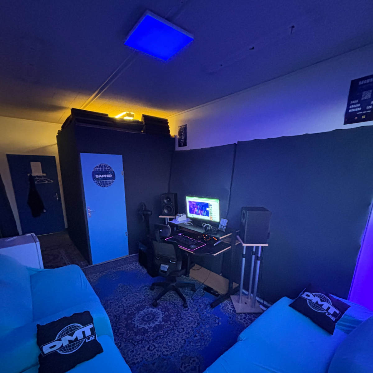 A cozy music studio featuring a computer setup with monitors, mixing equipment, and blue lighting, surrounded by dark walls and a comfortable seating area.