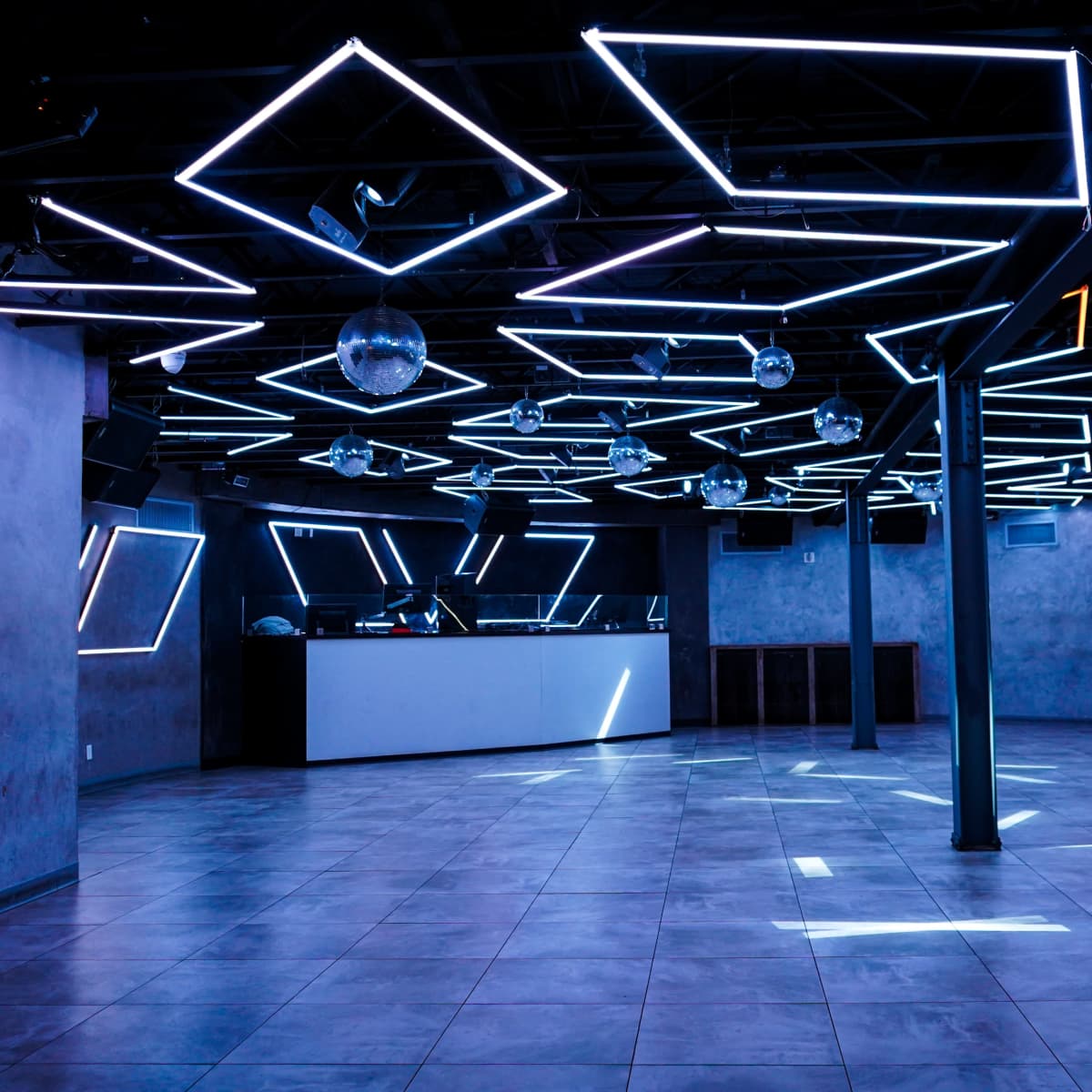 An empty nightclub interior with geometric neon light fixtures on the ceiling and a white bar in the background.