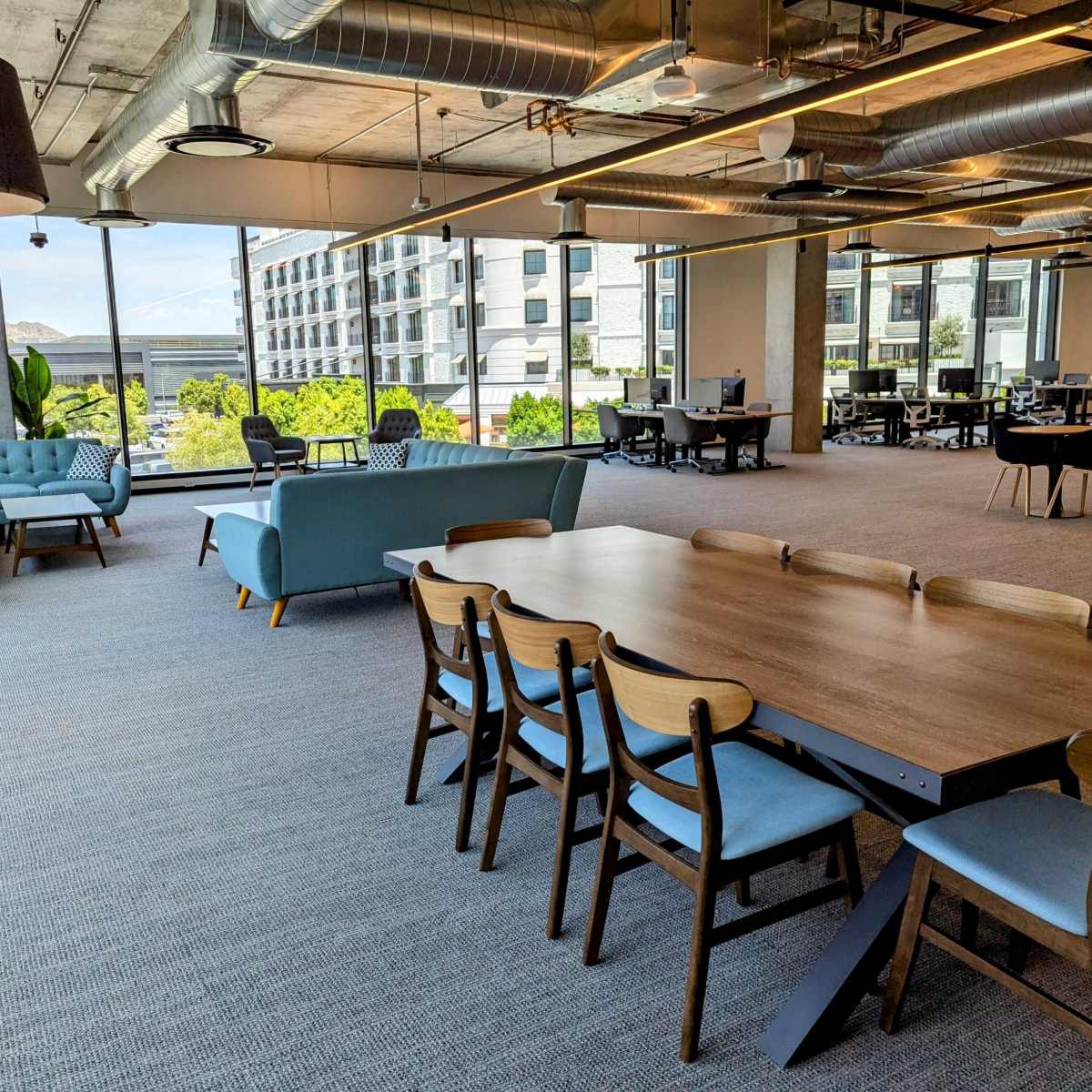 An open office space features a large wooden table surrounded by chairs, with couches and a view of outdoor greenery in the background.