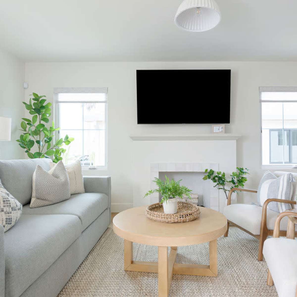 A modern living room with a light gray sofa, two armchairs, a round wooden coffee table, and a large television mounted above a white fireplace.