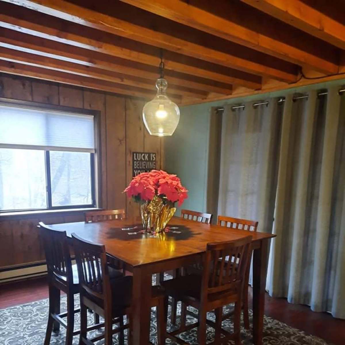 A wooden dining table surrounded by high-backed chairs is set under a pendant light in a room with wooden walls and a large window.