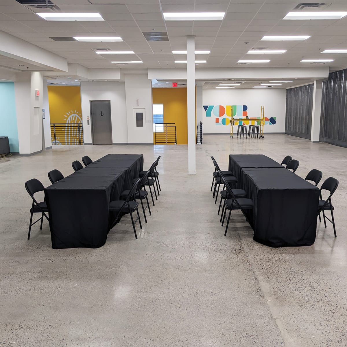 A spacious indoor area with two long tables covered in black tablecloths, surrounded by black chairs, and colorful wall art in the background.
