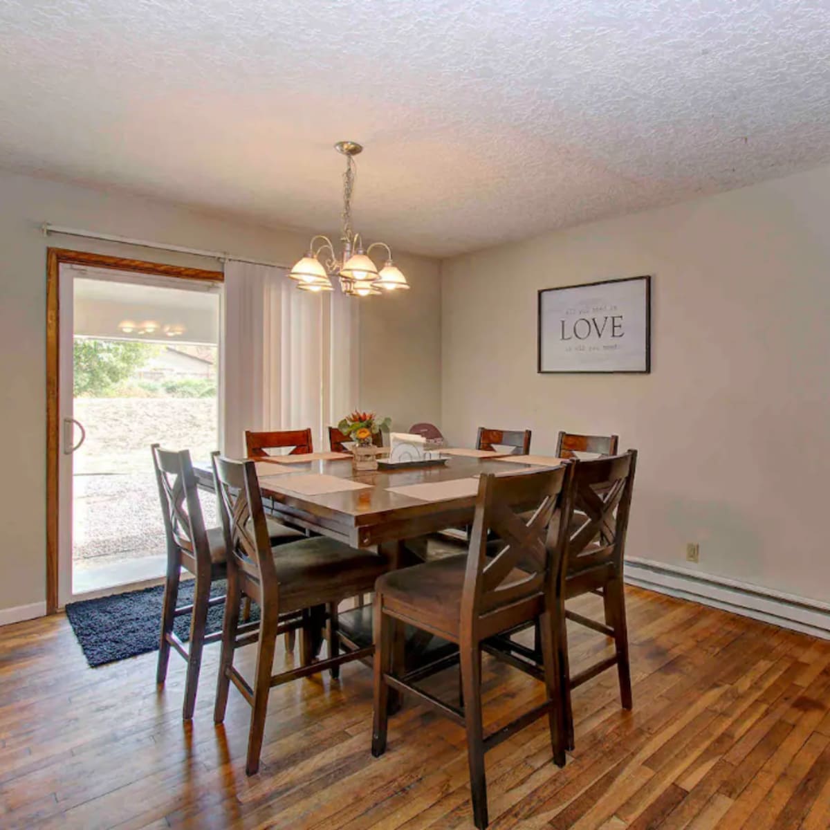 A dining area with a wooden table surrounded by eight chairs, accompanied by a chandelier and a sliding door leading outside.
