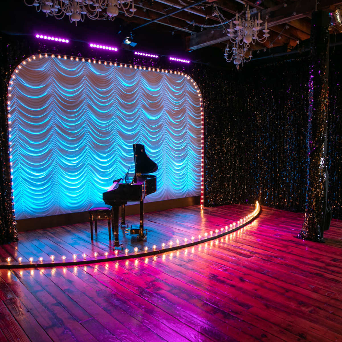 A performance stage with a grand piano, draped in soft blue fabric and surrounded by lights, located in a venue featuring a sign that reads "COTTON MILL LIVE."