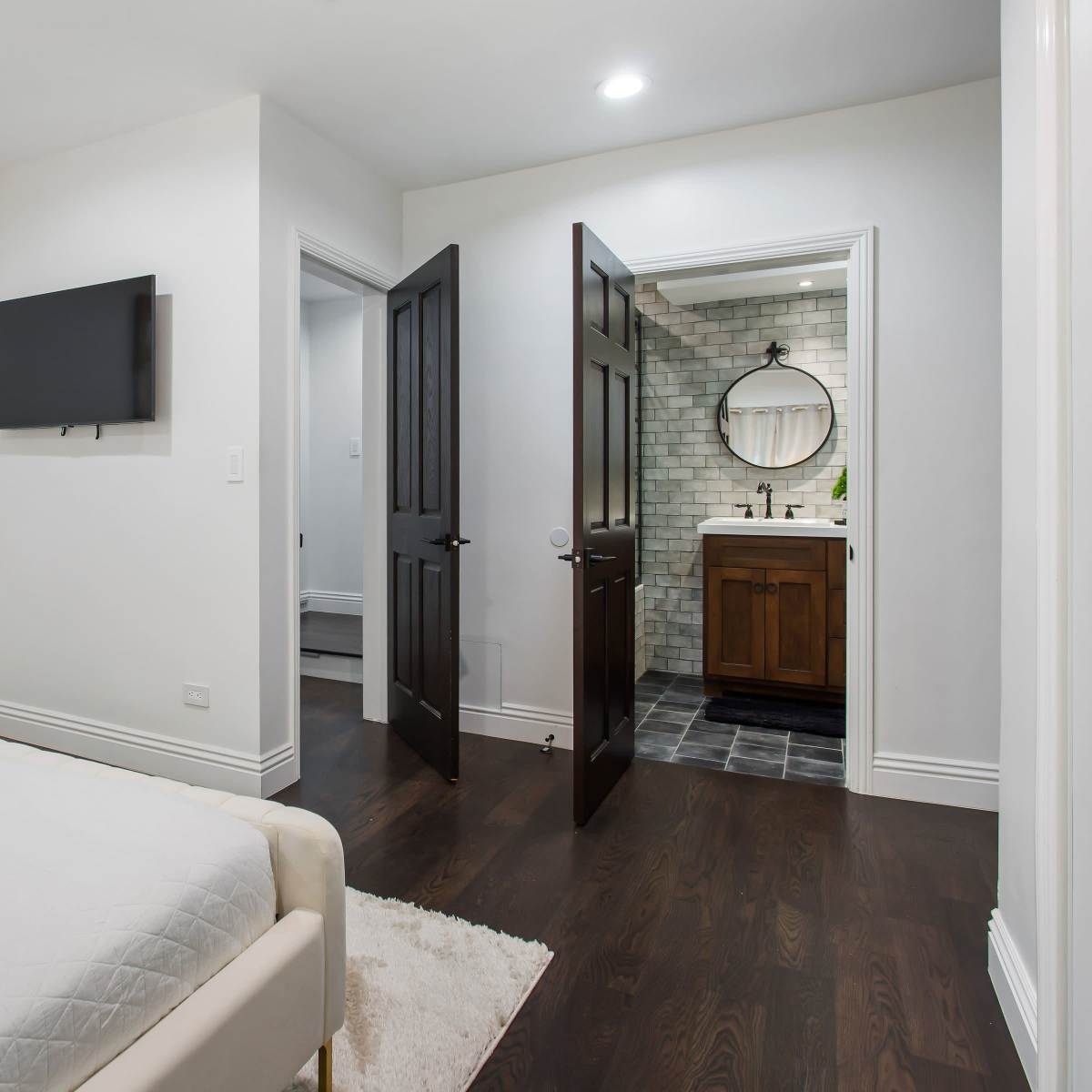 A neatly arranged bedroom features a white bed with a towel, and a doorway leading to a bathroom with a circular mirror and wooden vanity.