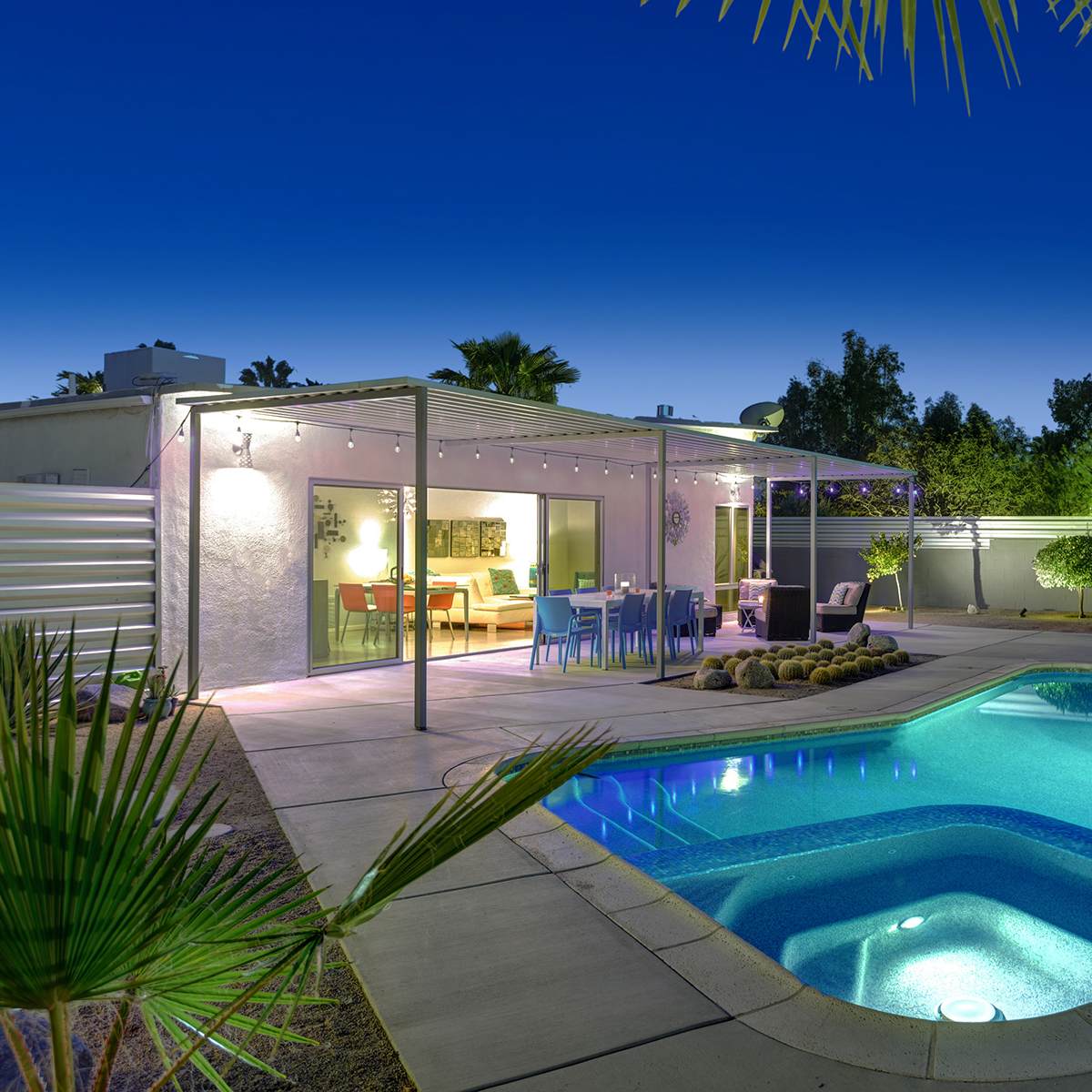 The scene features a modern backyard with a illuminated pool, a covered patio with dining furniture, and surrounding greenery under a clear evening sky.
