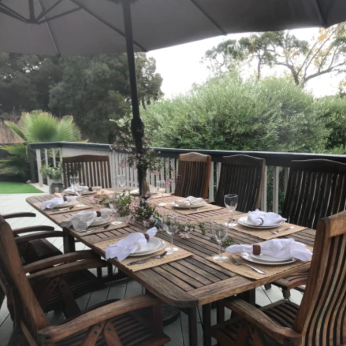 A wooden outdoor dining table is set with plates and glasses under a large umbrella on a deck surrounded by greenery.