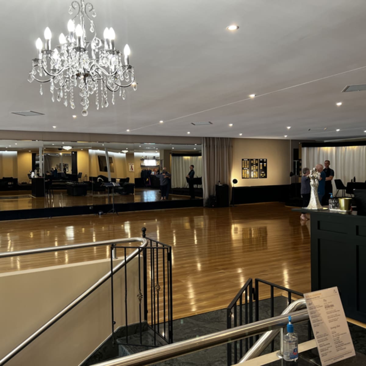 A spacious dance studio features polished wooden floors, a large mirror, and a chandelier overhead, with people dancing and socializing.