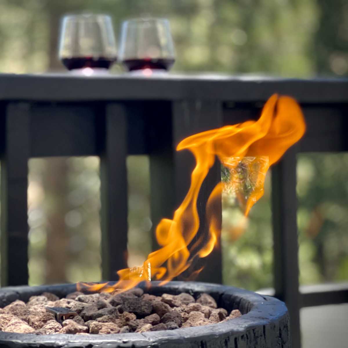 A lit fire pit with flames rising above a bed of rocks is positioned on a deck, with two glasses of red wine in the background.