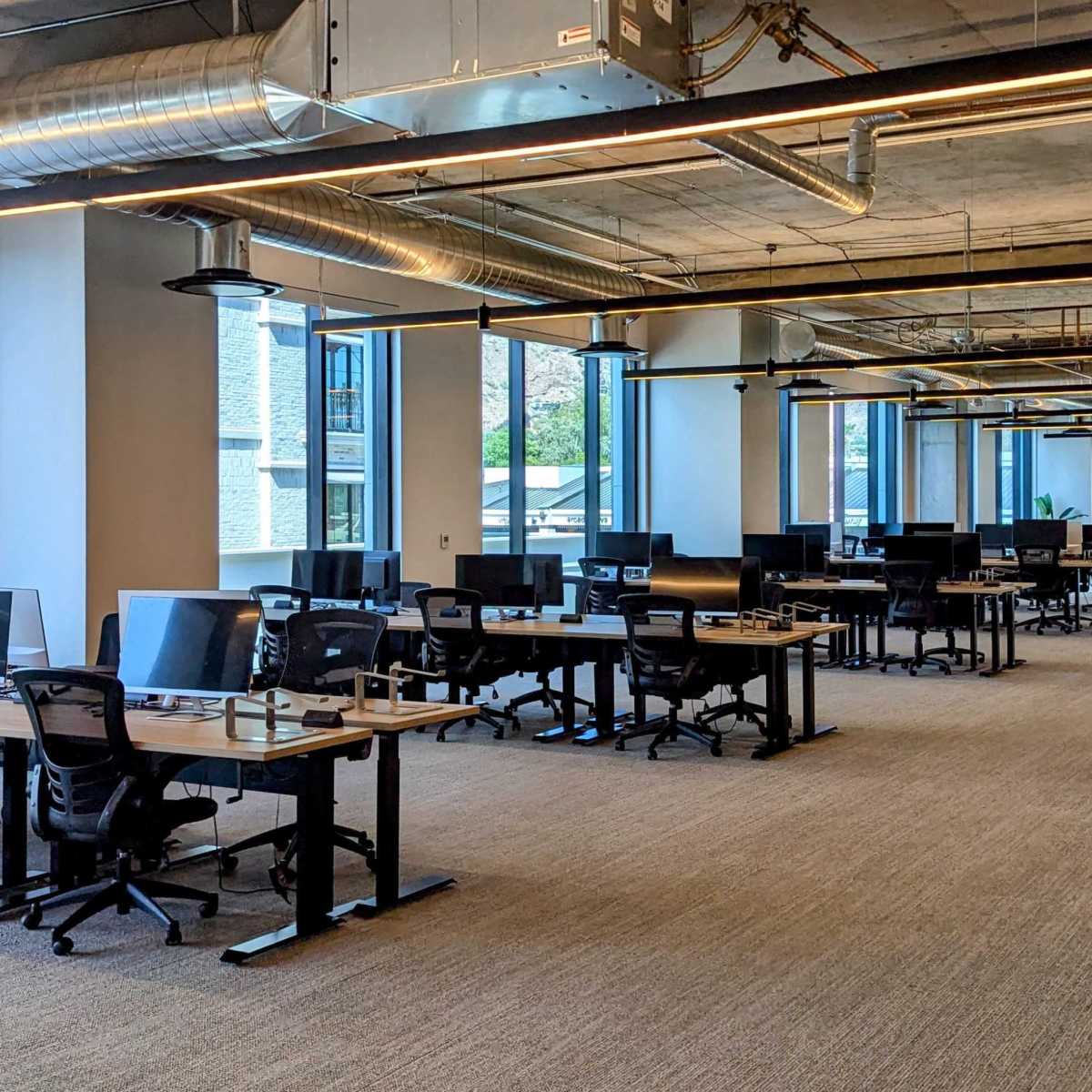 A modern office space with rows of desks and computers, large windows letting in natural light, and a neutral carpeted floor.