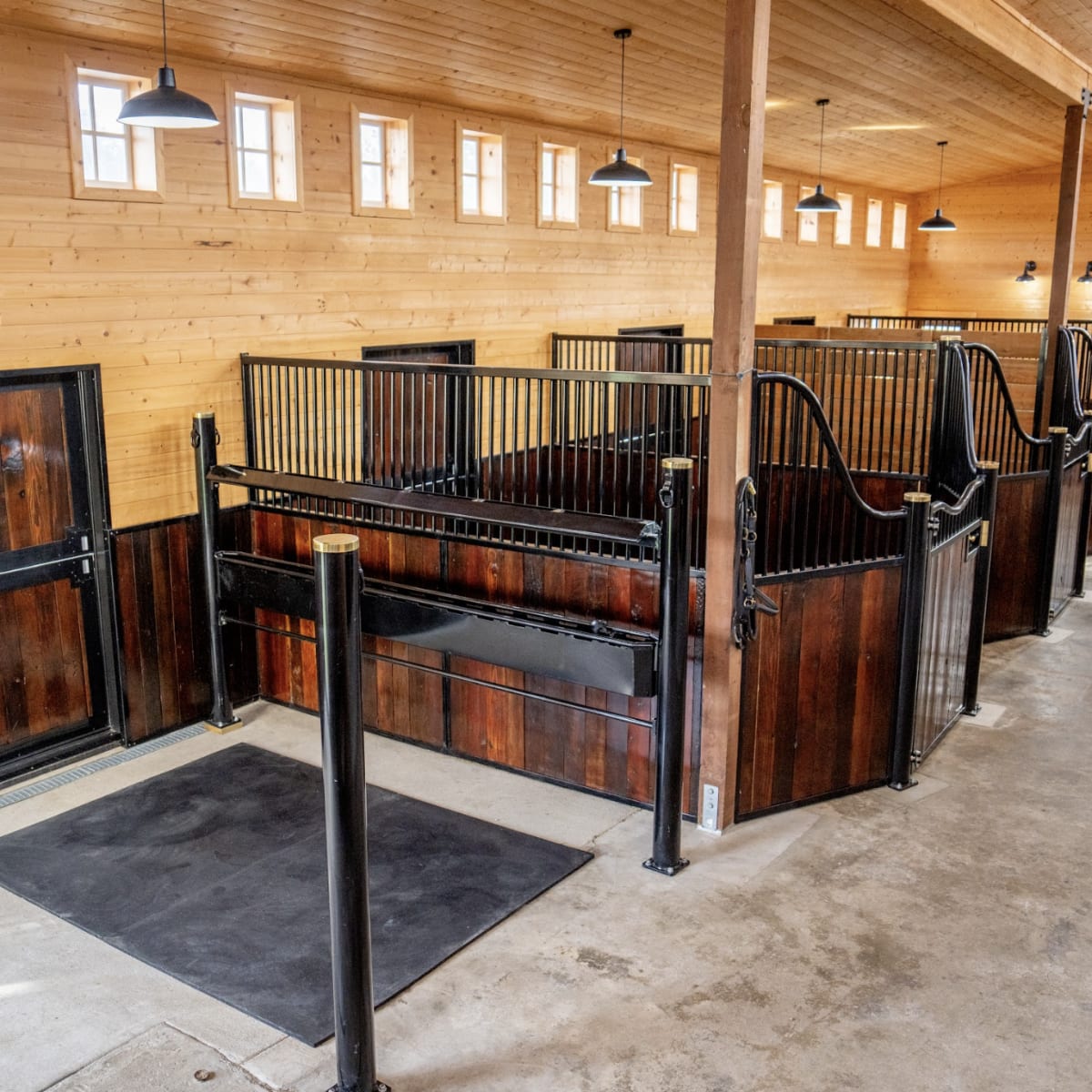 A spacious stable interior with wooden walls, several horse stalls on each side, and a concrete floor.