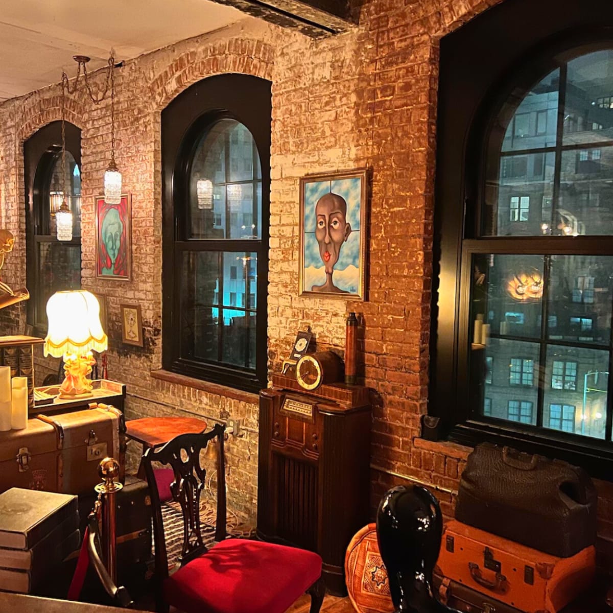 A cozy interior filled with vintage furniture and decor, including a lamp, books, and artwork, alongside large arched windows revealing a cityscape at night.