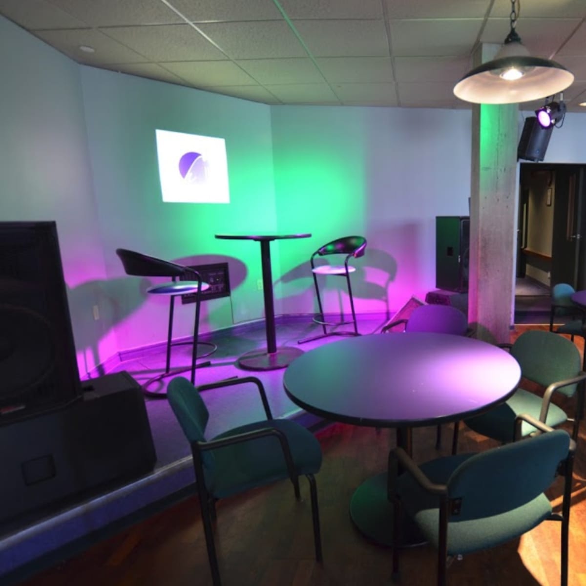 A small lounge area featuring round tables and chairs, a stage with a colorful light setup, and a projector screen against the wall.