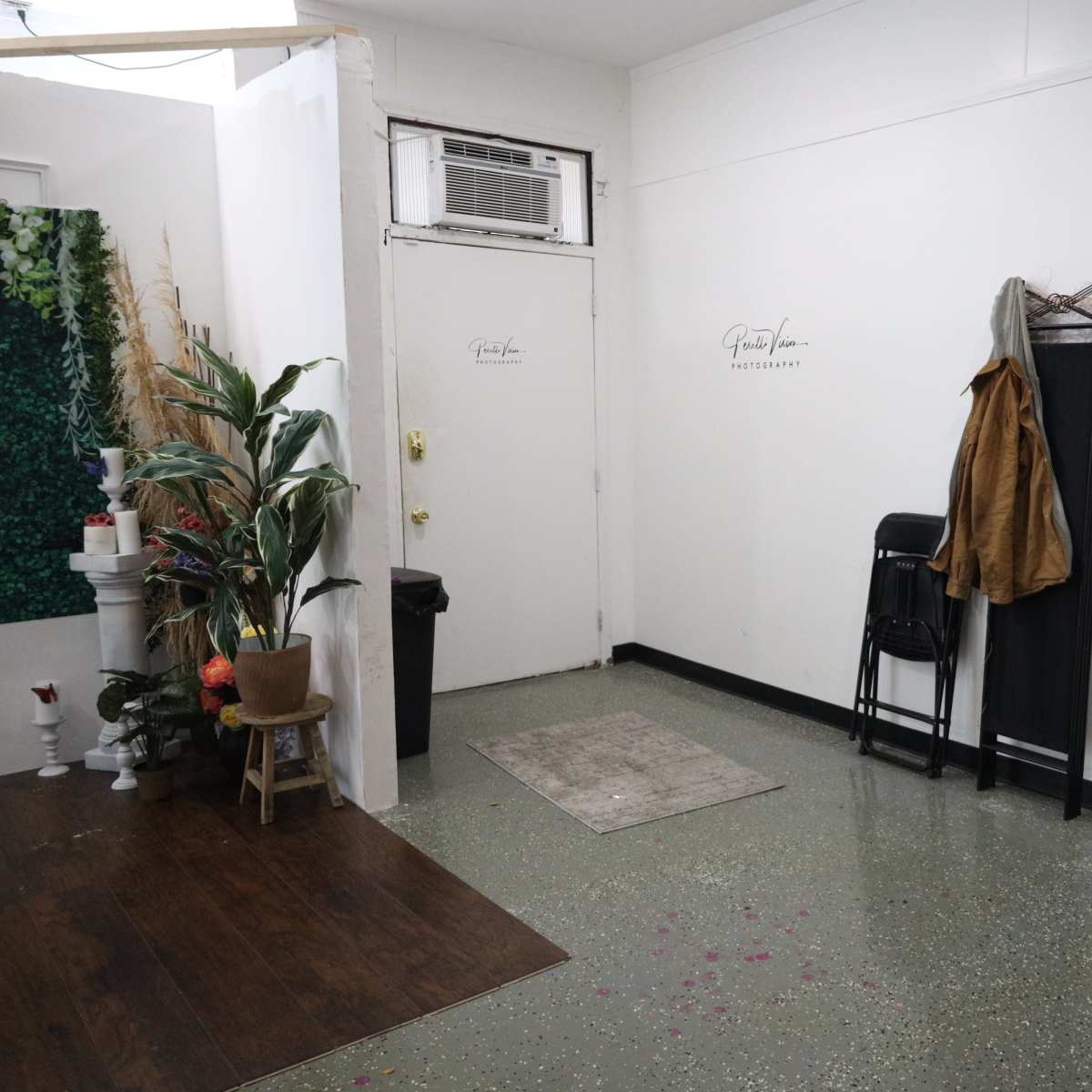 A small, sparsely furnished room with a few chairs, a coat hanging on a rack, and a plant next to a wall adorned with greenery.