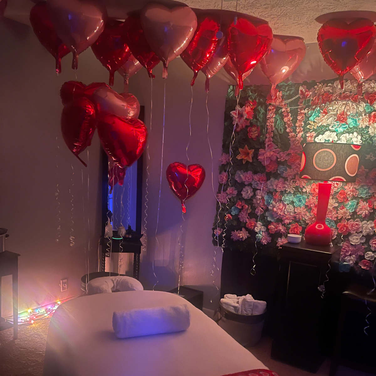 The room is decorated with heart-shaped balloons and floral wall decor, featuring a massage table covered with a blanket and a rolled towel.