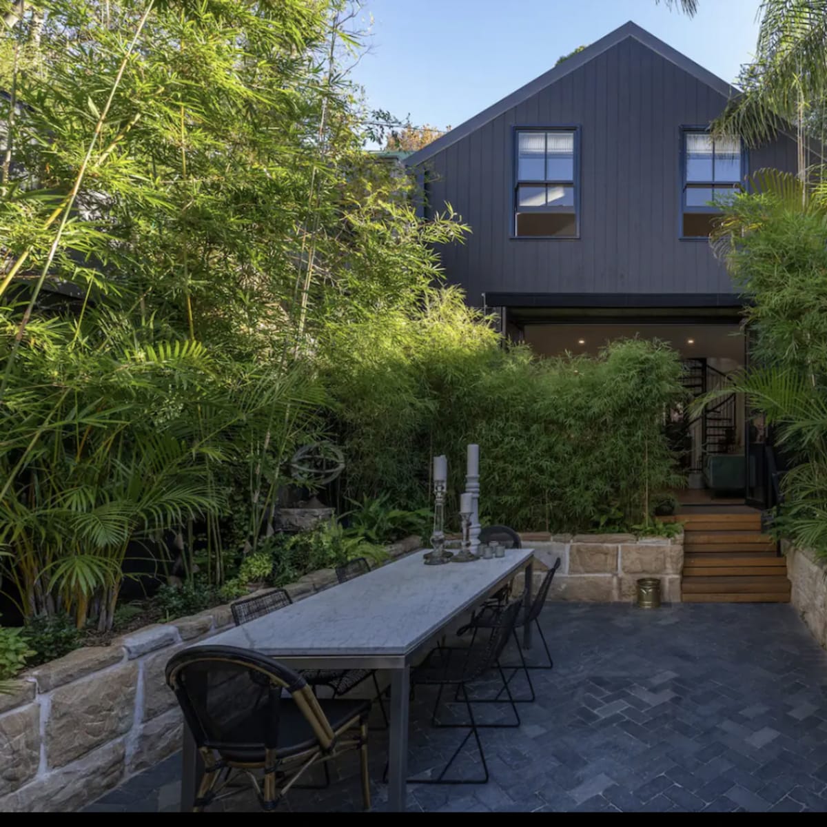 A stone pathway leads to a modern black house surrounded by vibrant greenery and a long dining table in a tranquil garden setting.