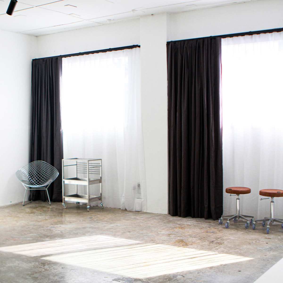 A bright, minimalist room with two large windows, dark curtains, a wire chair, a metal cart, and two stools.