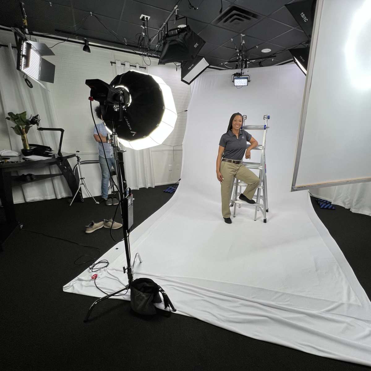 A woman sits on a ladder in a studio setup with lighting equipment and a backdrop.