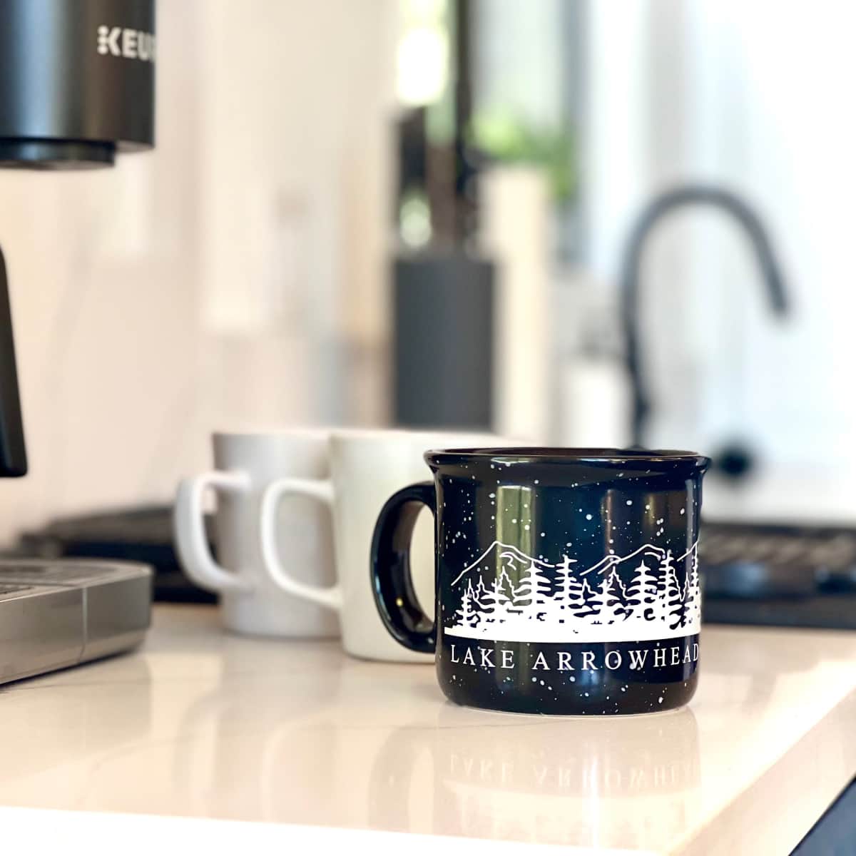 A coffee maker sits on a countertop next to several mugs, including a blue one with a "Lake Arrowhead" design.