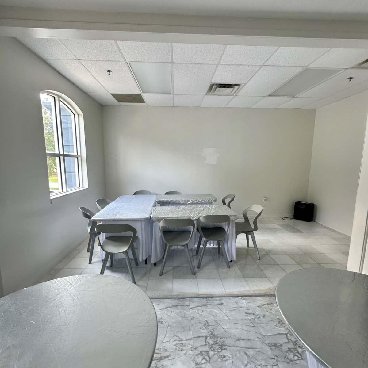 A sparsely furnished room features a table surrounded by chairs, with two small round tables in the foreground and a large window letting in natural light.