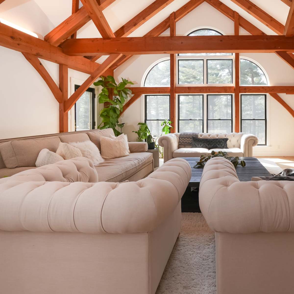A spacious living room features two tufted sofas facing a large window with a view of the outdoors, surrounded by exposed wooden beams and greenery.