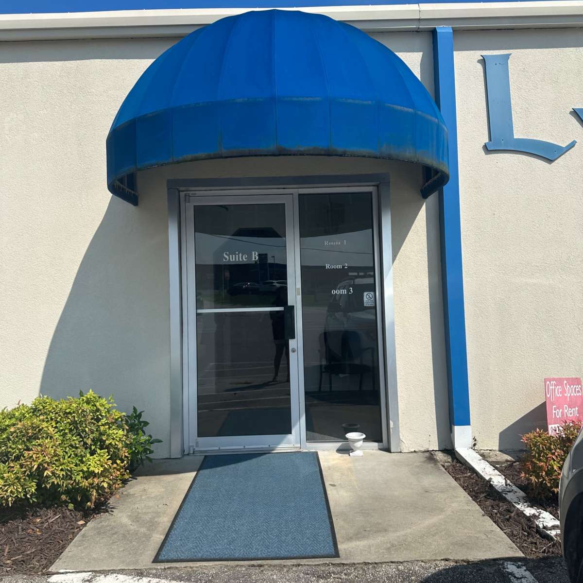 The entrance of a commercial building features a blue awning above double glass doors, with a blue mat leading to the entrance.