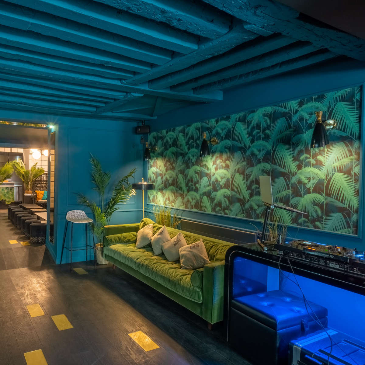 A vibrant blue room with a green velvet sofa, palm leaf wallpaper, and wooden beams on the ceiling, leading to an illuminated entryway.