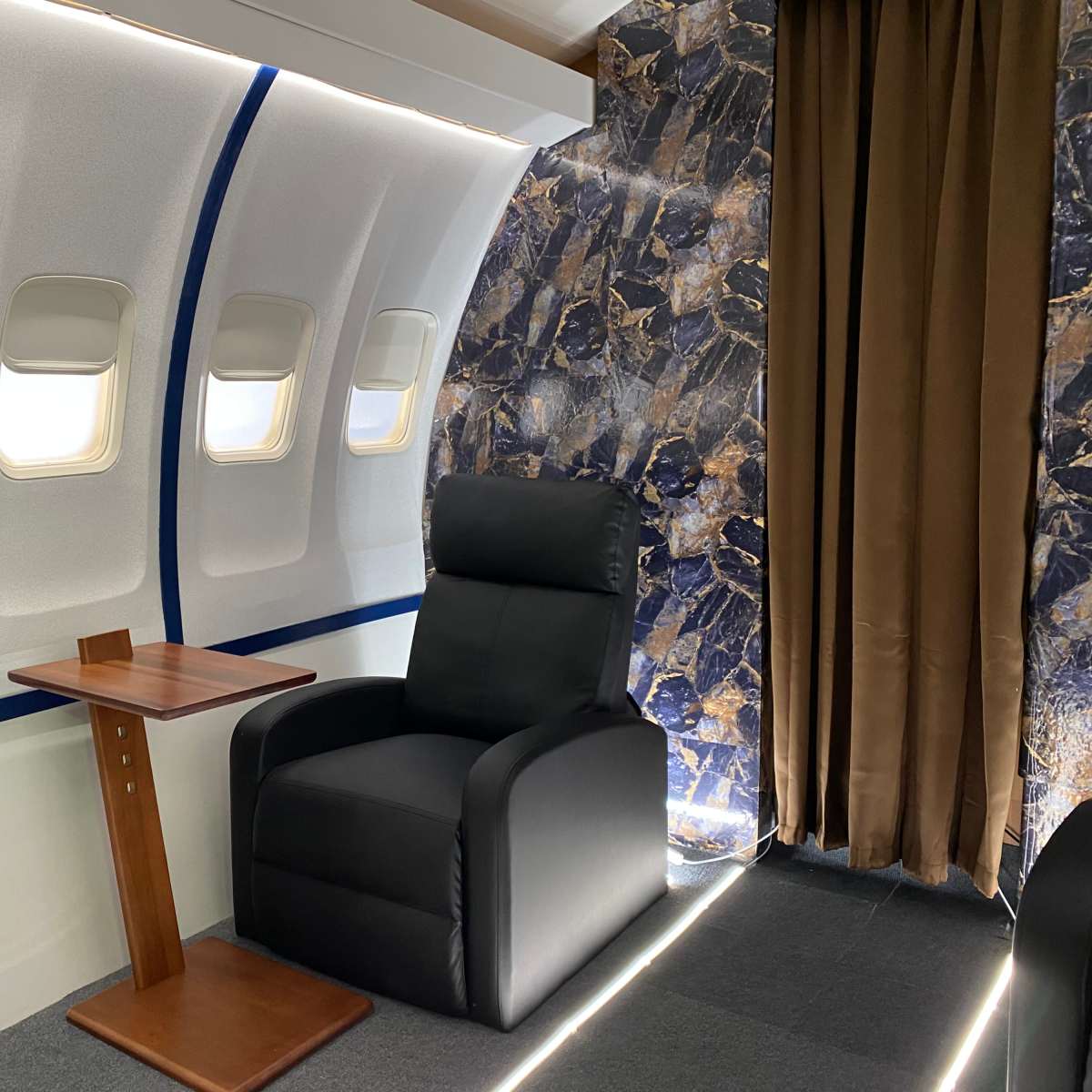 A luxurious interior of an aircraft cabin with a black armchair, a small wooden side table, and large windows.