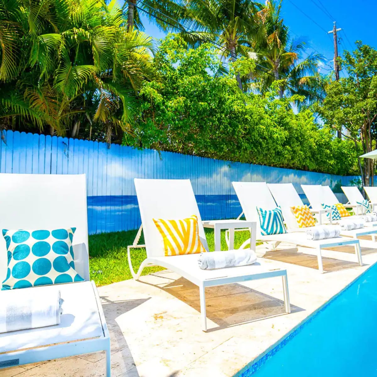 A bright poolside area with white lounge chairs arranged neatly beside a clear blue pool, surrounded by greenery and a mural of the ocean.