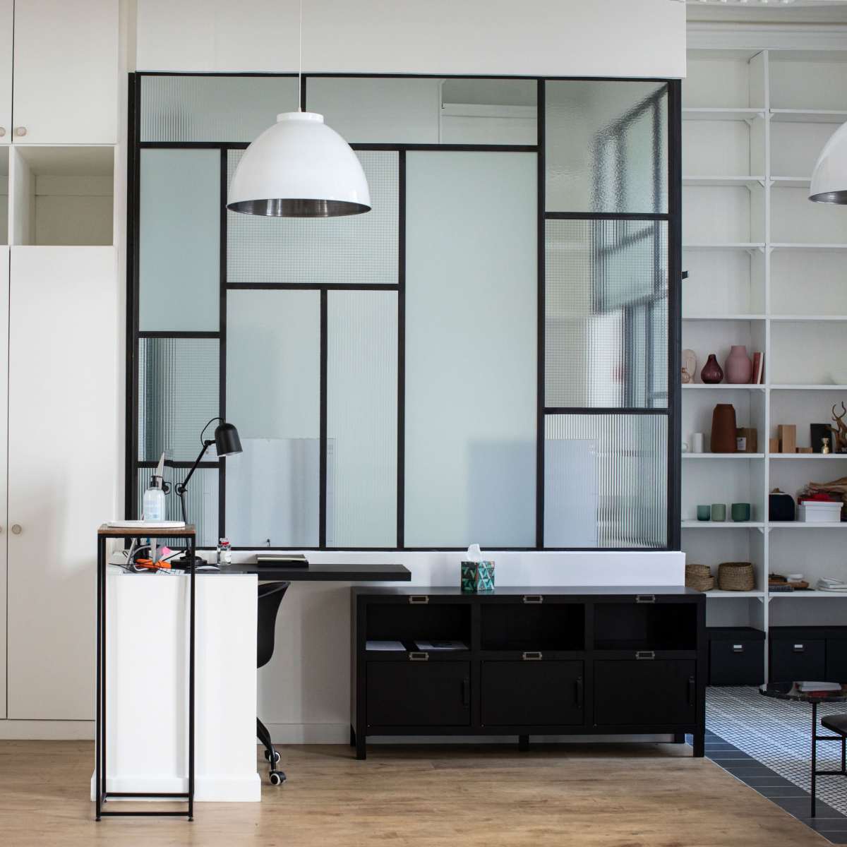 A modern office interior featuring a black desk and a partition with geometric glass panels, surrounded by white cabinetry and shelving.