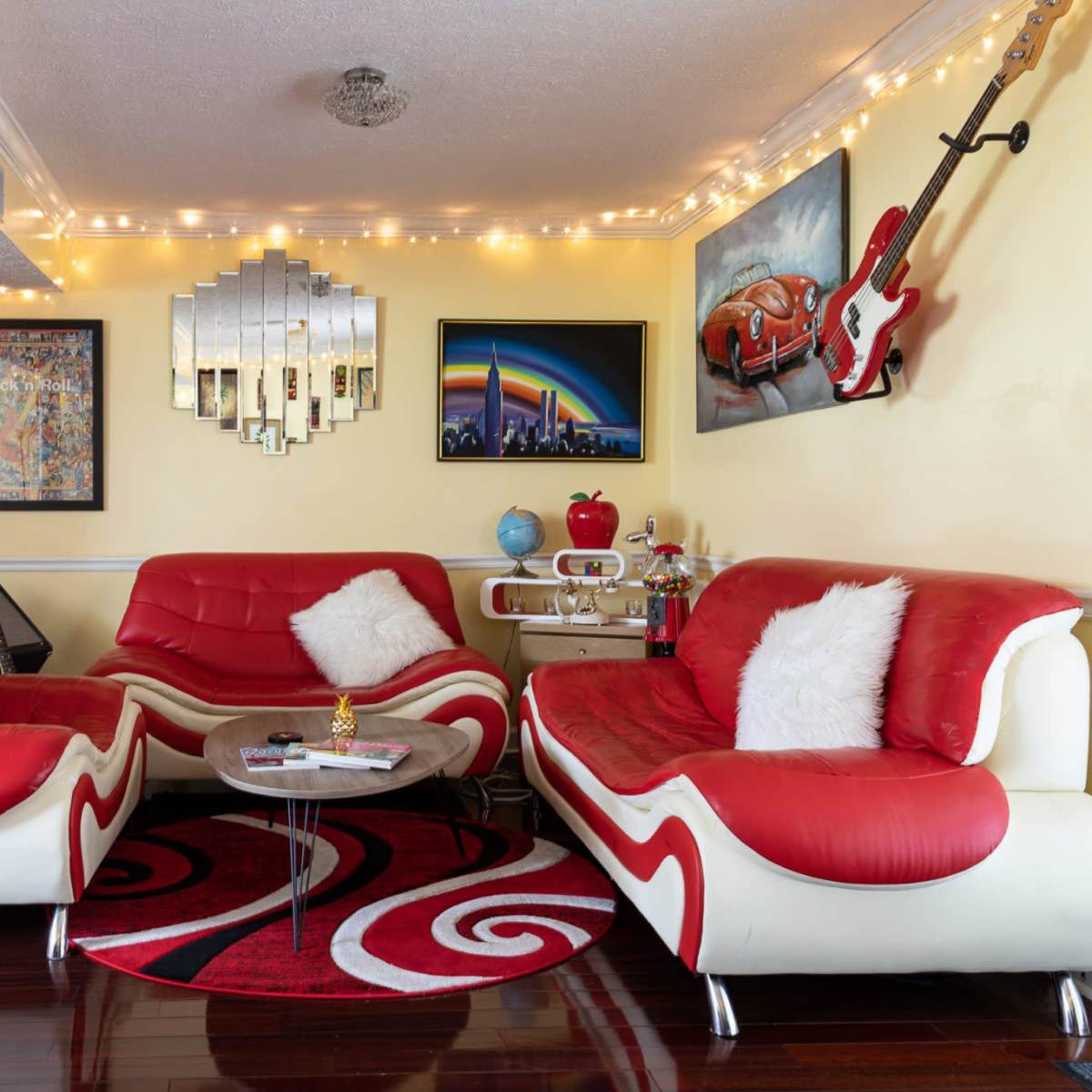 A vibrant living room with red and white furniture, a round rug, framed artwork on the walls, and decorative string lights.