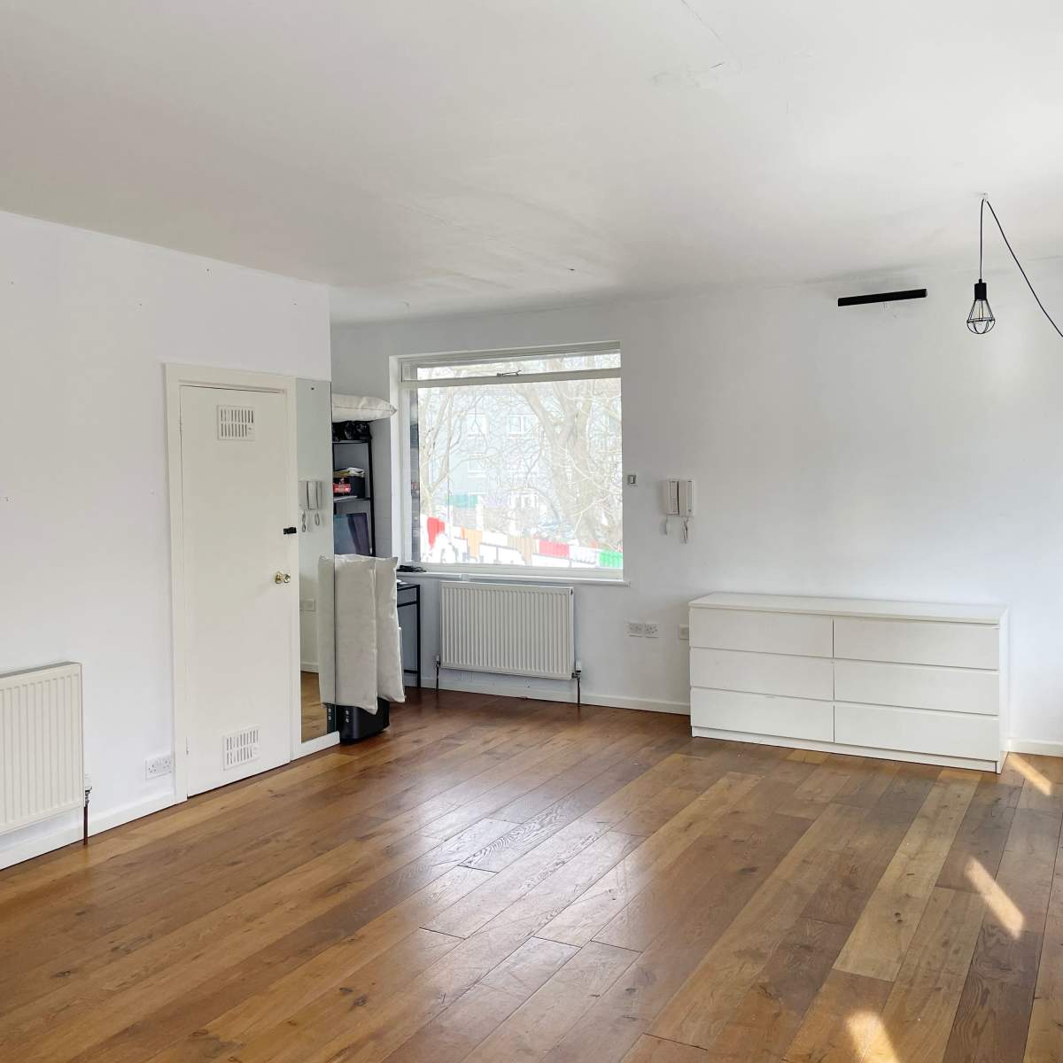 A spacious room with wooden flooring, a large window, a white dresser, and a light fixture hanging from the ceiling.