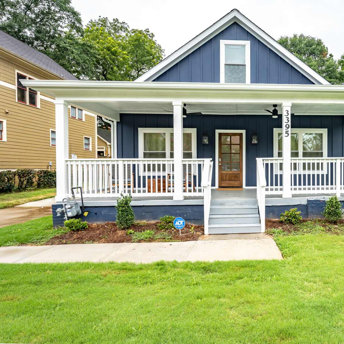 A blue two-story house with a front porch and white railings is set on a grassy lawn with landscaping.