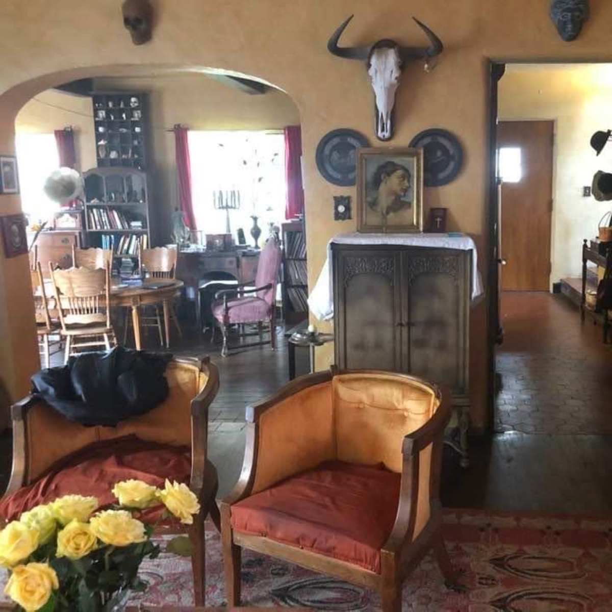 A warmly decorated room featuring two orange armchairs, a wooden dining table with chairs, and a hallway leading to another room, adorned with various wall decorations and a mounted animal skull.