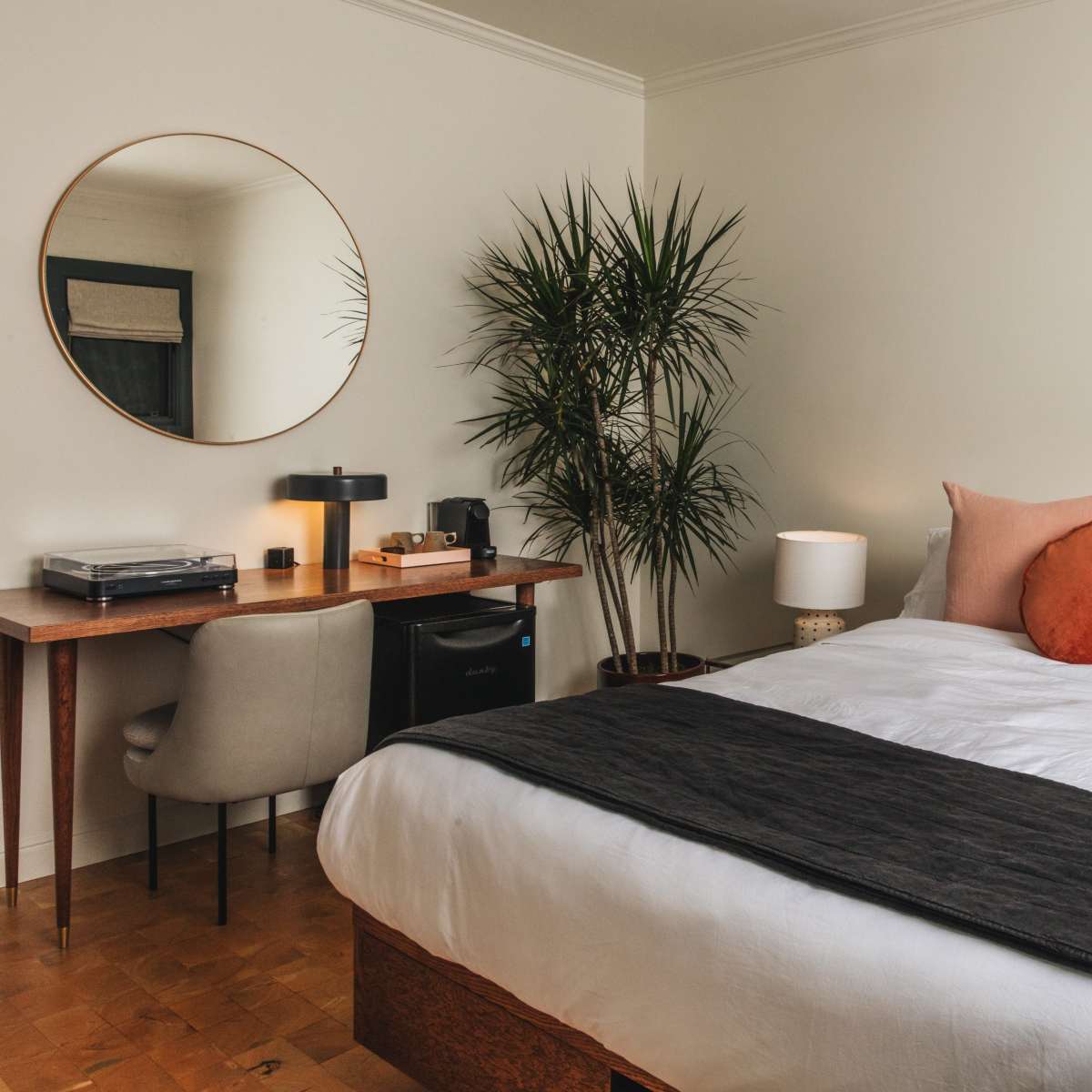 A neatly arranged bedroom features a large bed with decorative pillows, a desk with a lamp and a record player, and a potted plant.