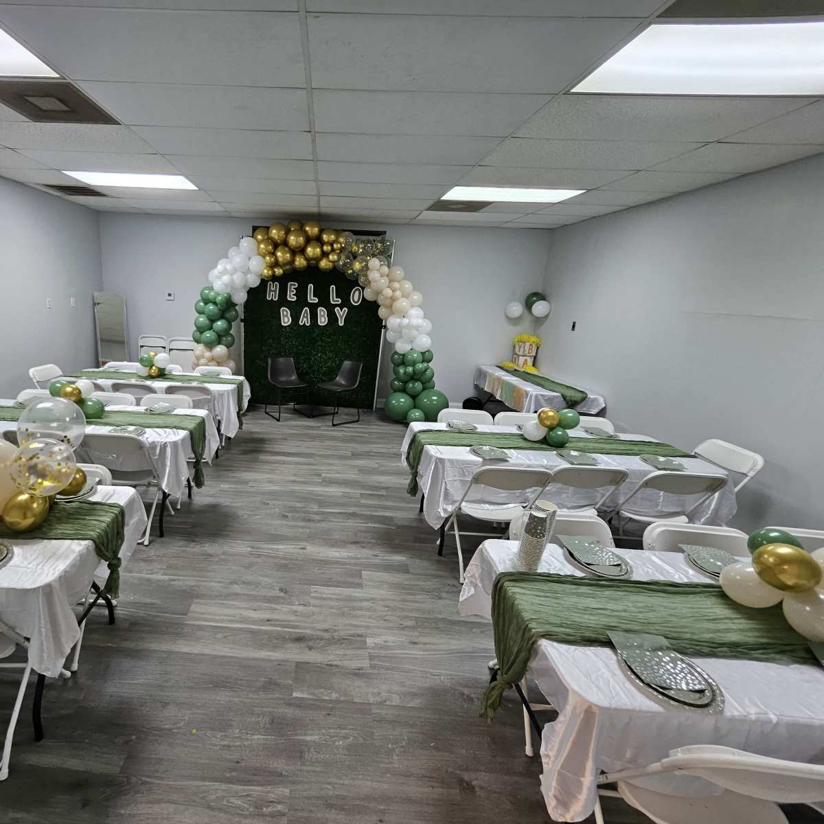 A decorated room for a baby shower, featuring several tables with white tablecloths, green and gold balloons, and a backdrop that reads "HELLO BABY."
