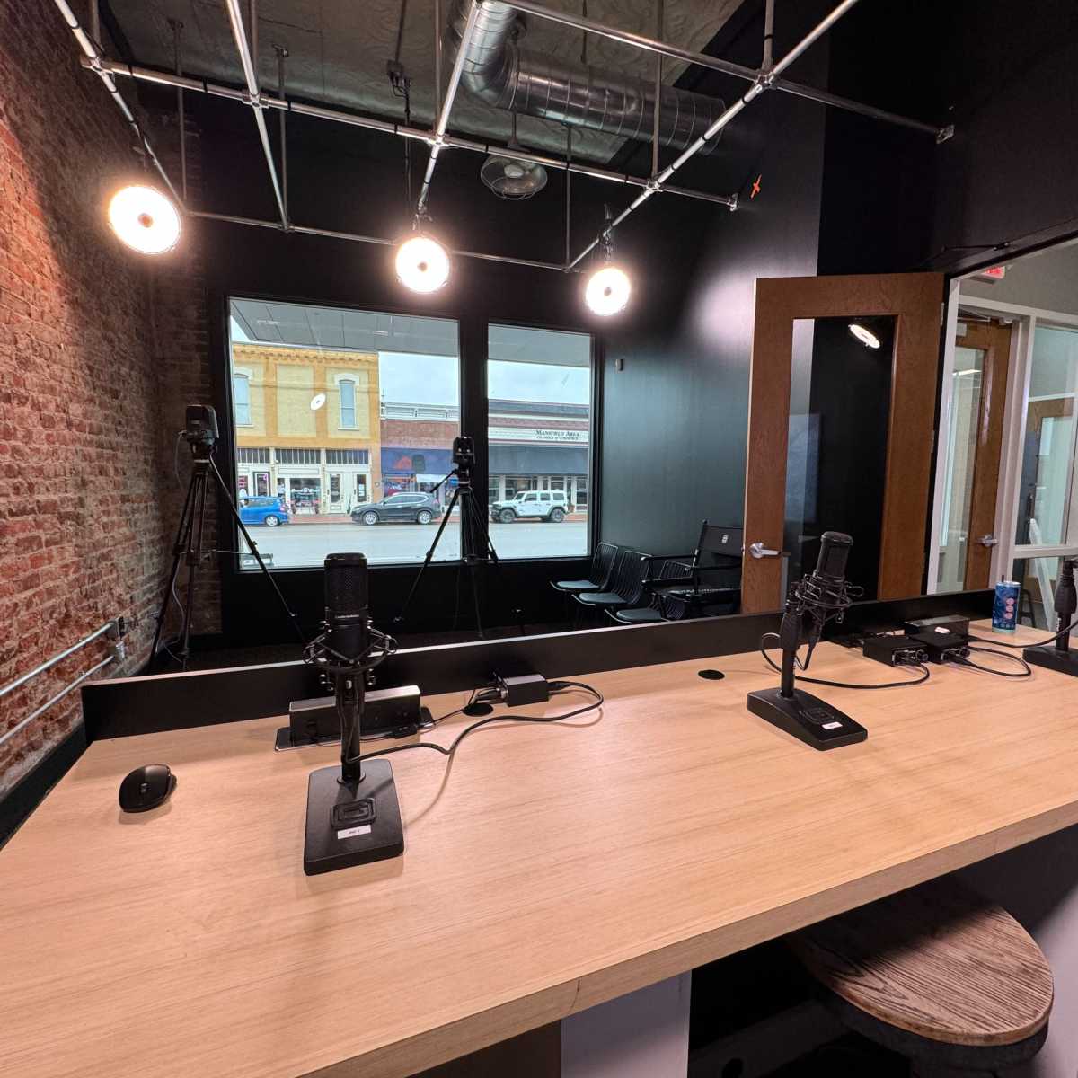 A podcast recording studio featuring a wooden table with microphones, chairs, and large windows overlooking a street.