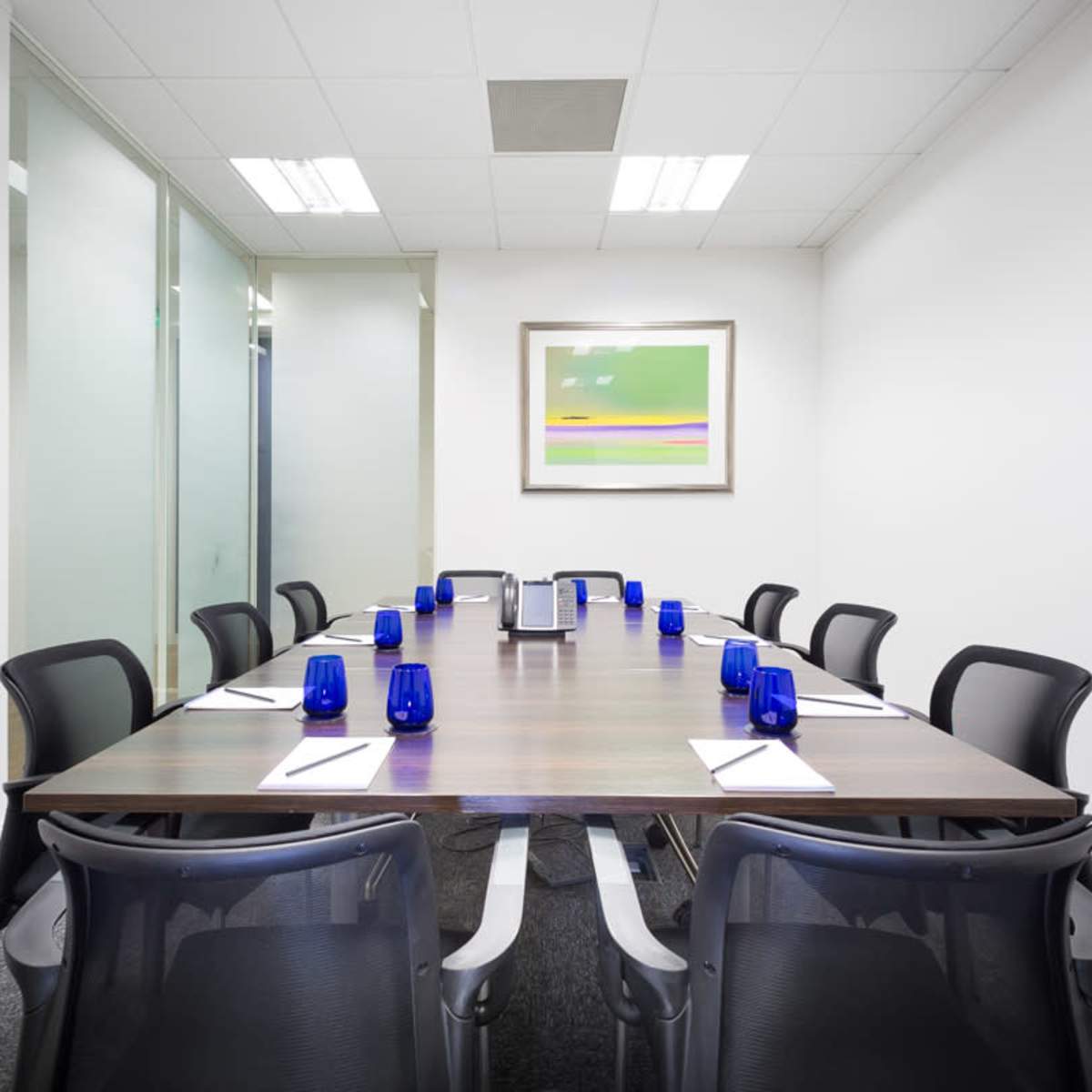 A long wooden conference table is set with blue glasses and notepads, surrounded by black chairs in a brightly lit meeting room.