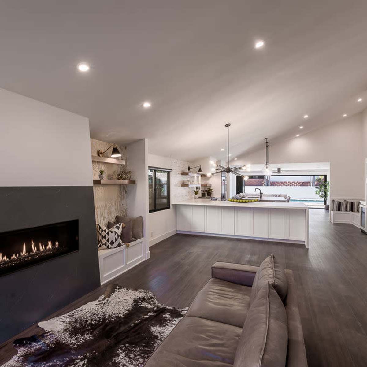 A modern living room with a fireplace, a large open kitchen, and an area with neutral furniture and hardwood flooring.