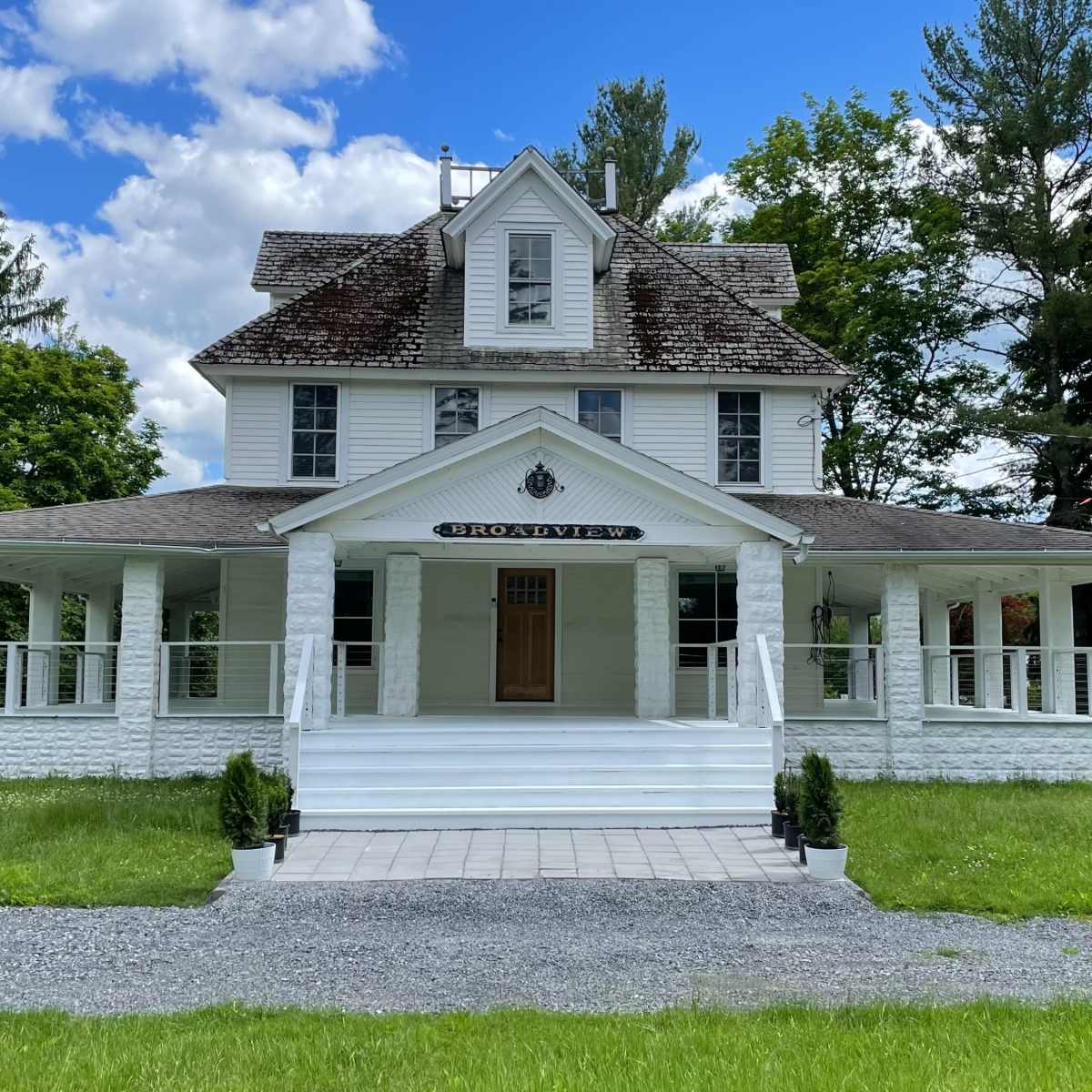 A large, white house with a wraparound porch, set on a grassy area with a gravel driveway under a blue sky dotted with clouds.