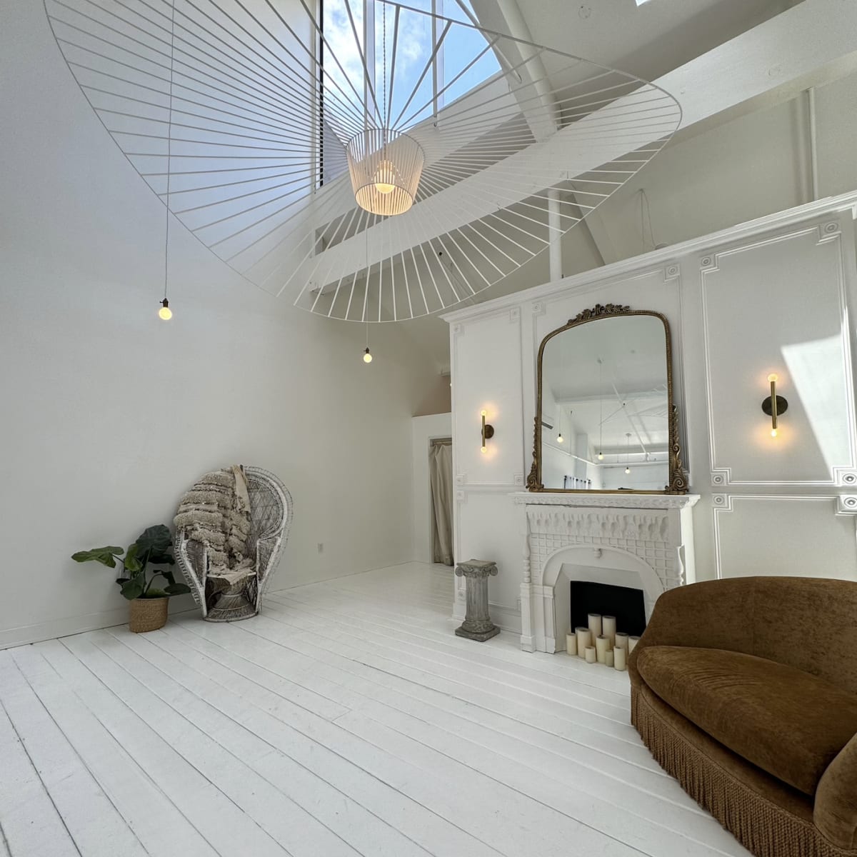 A spacious, minimalist room with a high ceiling, featuring a large mirror above a fireplace, a hanging light fixture, and a wicker chair surrounded by white walls and wooden flooring.