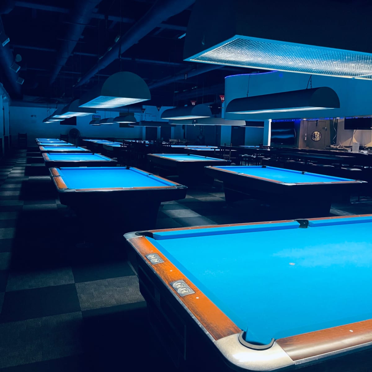 A dimly lit room filled with pool tables, each illuminated by overhead lights, casting a blue glow over the table surfaces.