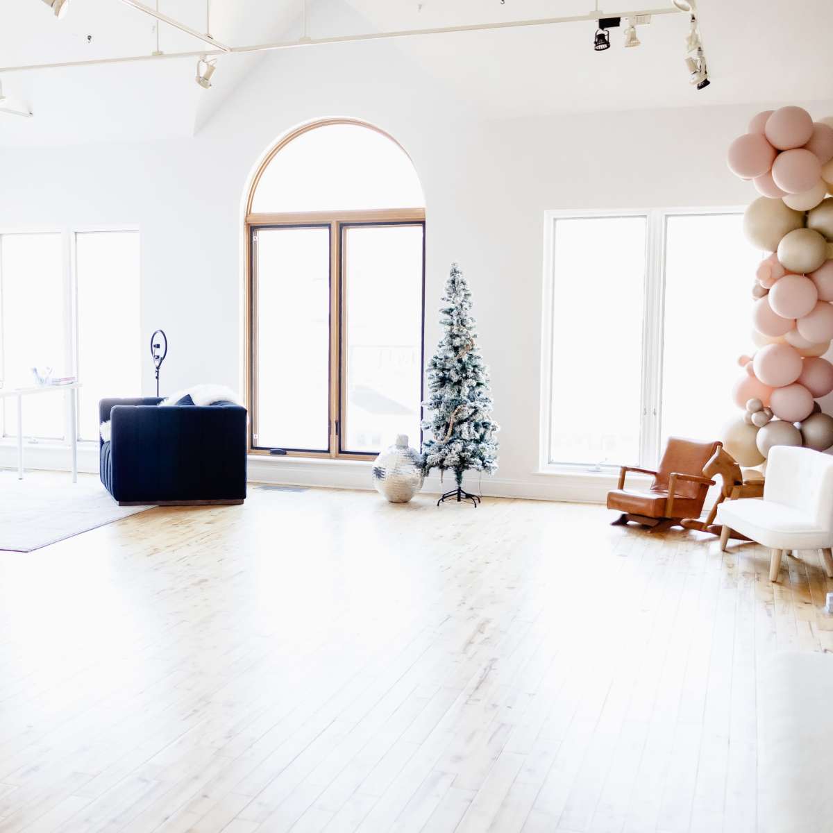A spacious, well-lit room with wooden flooring, featuring a Christmas tree, a desk with a chair, and a seating area with a couch and a rocking chair.
