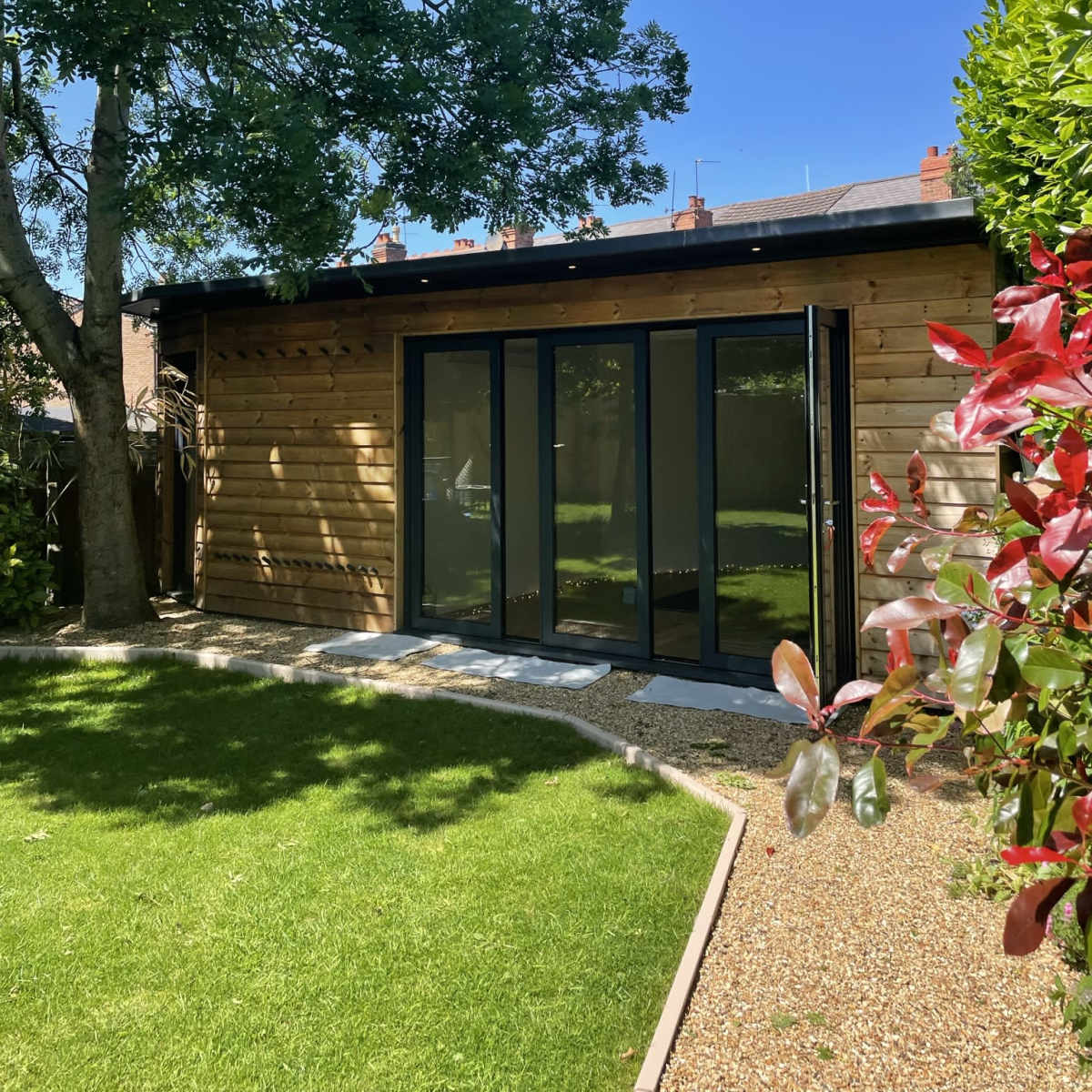 A wooden garden shed with large glass doors is situated in a green backyard surrounded by trees and shrubbery.