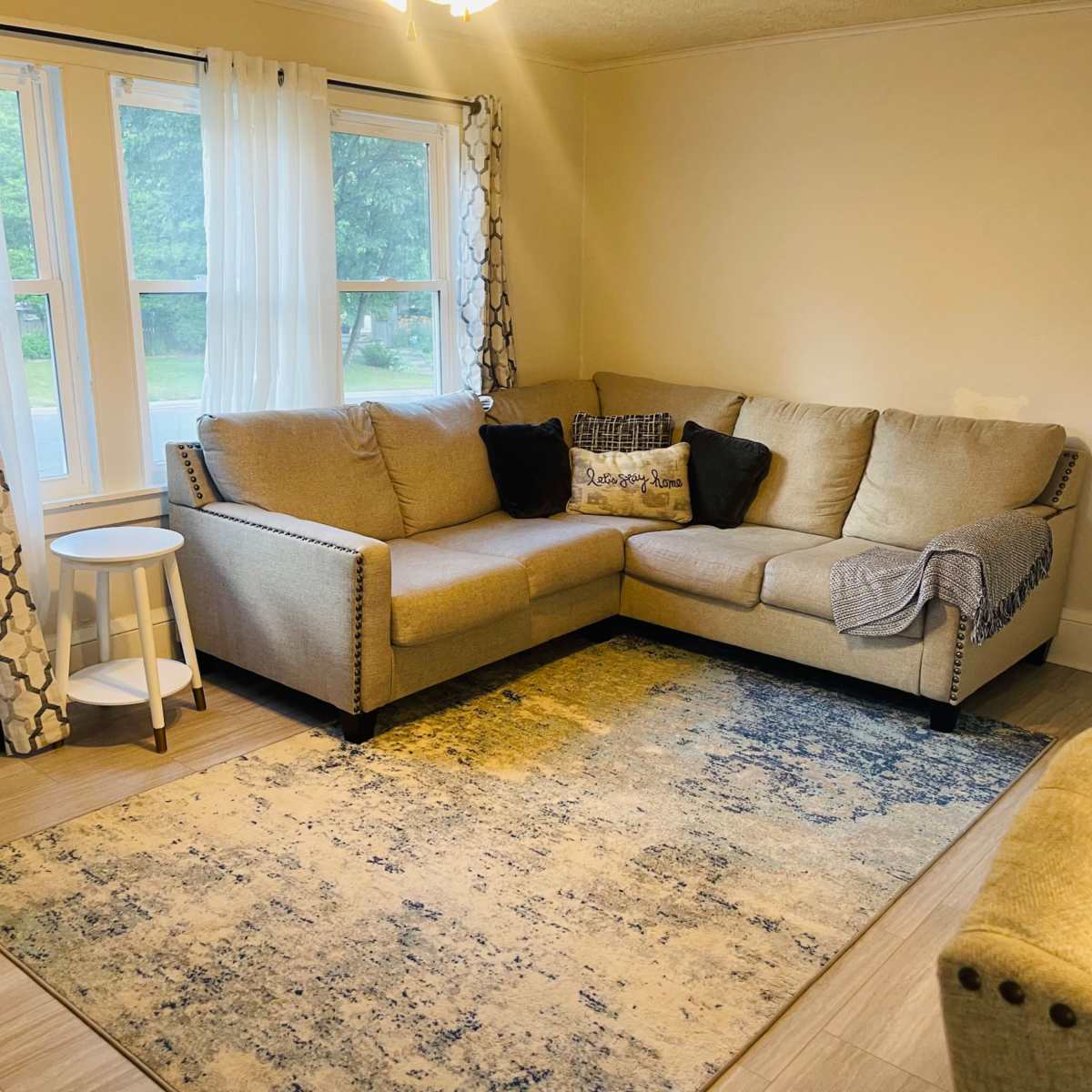 A cozy living room with a sectional sofa, a round side table, and a large area rug, all positioned beside two windows with sheer curtains.