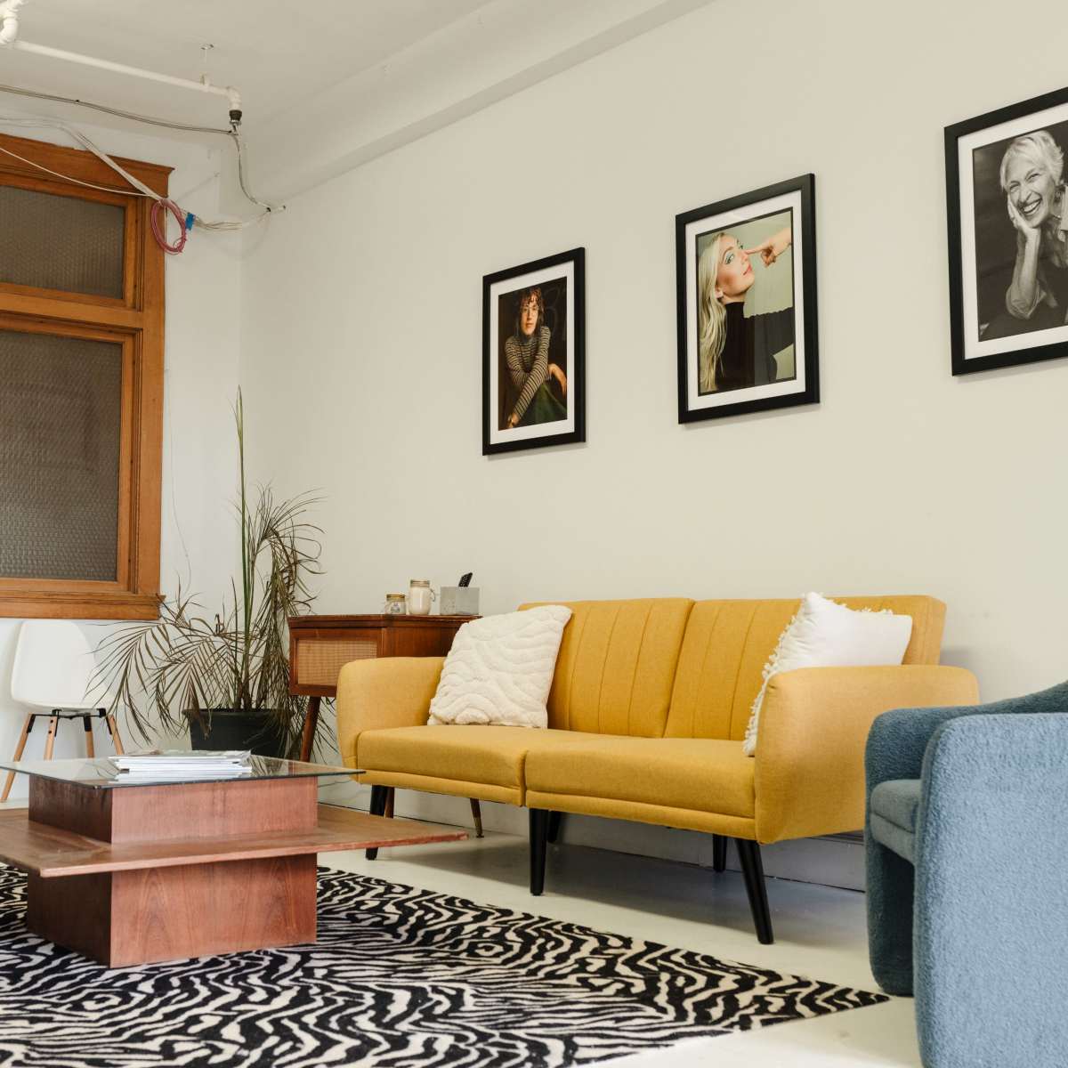 A modern seating area featuring a yellow couch and a blue chair, with a coffee table on a patterned rug and framed black-and-white photographs on the wall.
