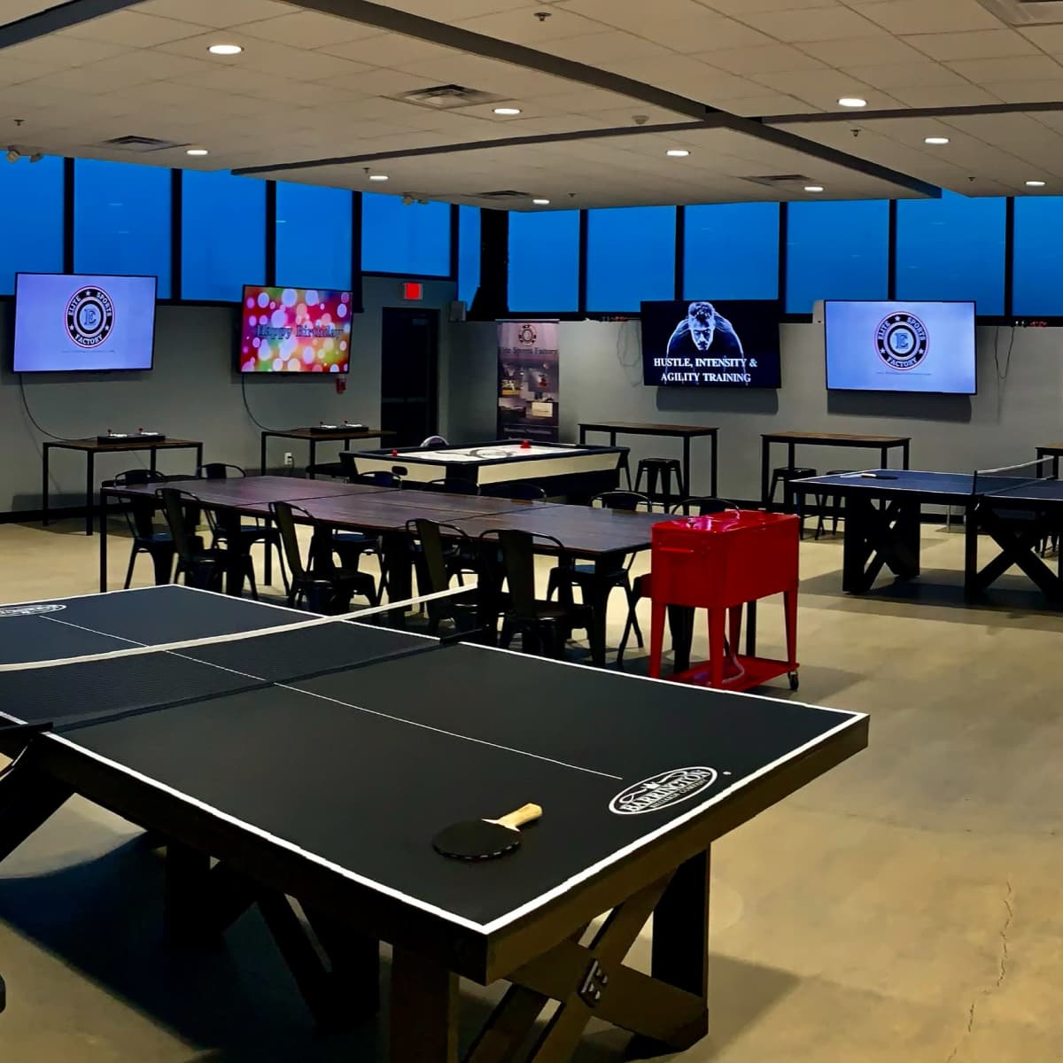A modern game room featuring multiple air hockey tables, foosball tables, and large screens mounted on the walls, with large windows displaying a view of the outdoors.