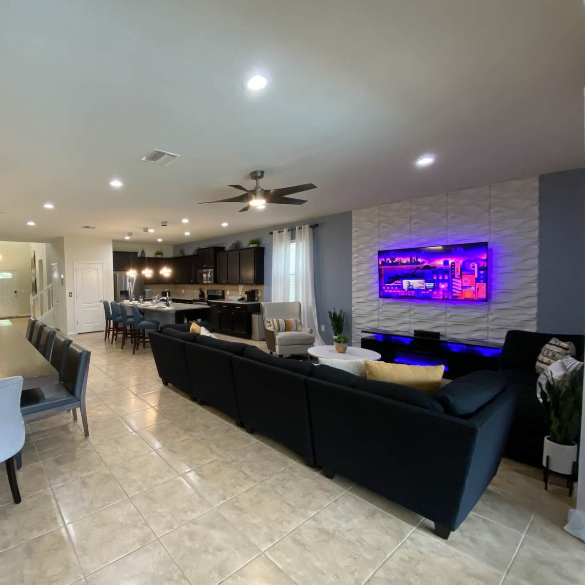 A modern open-concept living and dining area with a long dining table, a sectional sofa, and a kitchen visible in the background.